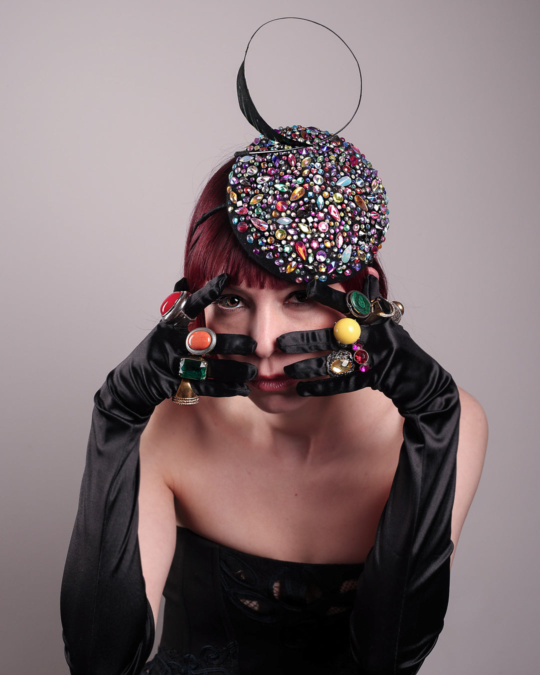 The model showcases the full  top of the Celestine pillbox fascinator. Its structured, button-shaped black base is drenched in a kaleidoscope of iridescent crystals—pink, aqua, violet, and gold, among others—each catching the light in motion. Crowned with a sweeping black quill that curves like a clef in mid-air. Designed by Merve Bayindir.