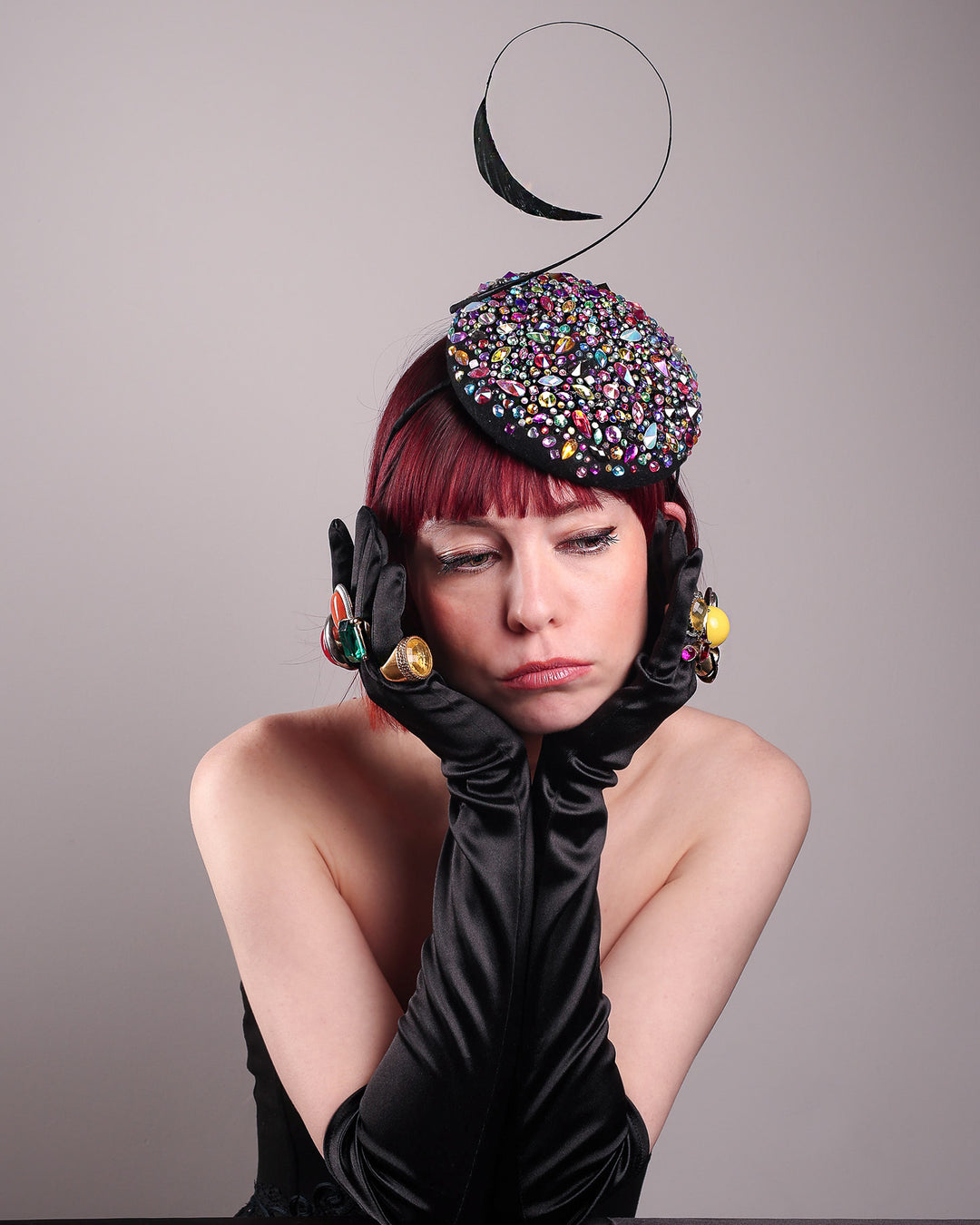 The model showcases the top front of the Celestine pillbox fascinator. Its structured, button-shaped black base is drenched in a kaleidoscope of iridescent crystals—pink, aqua, violet, and gold, among others—each catching the light in motion. Crowned with a sweeping black quill that curves like a clef in mid-air. Designed by Merve Bayindir.