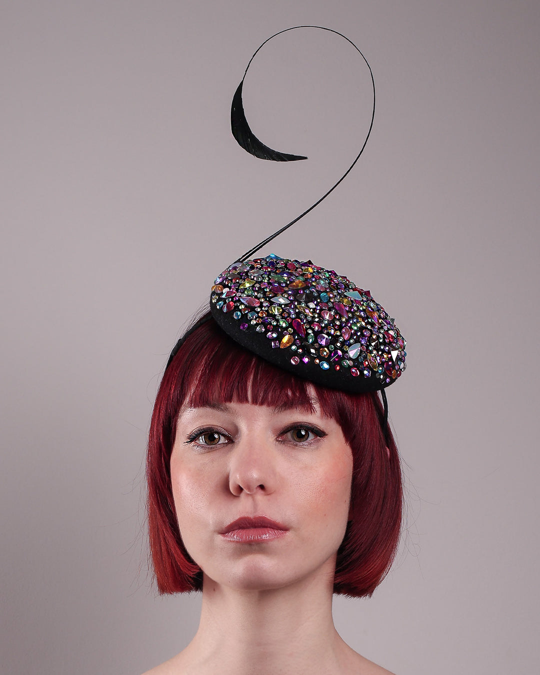 The model showcases the front close up of the Celestine pillbox fascinator. Its structured, button-shaped black base is drenched in a kaleidoscope of iridescent crystals—pink, aqua, violet, and gold, among others—each catching the light in motion. Crowned with a sweeping black quill that curves like a clef in mid-air. Designed by Merve Bayindir.