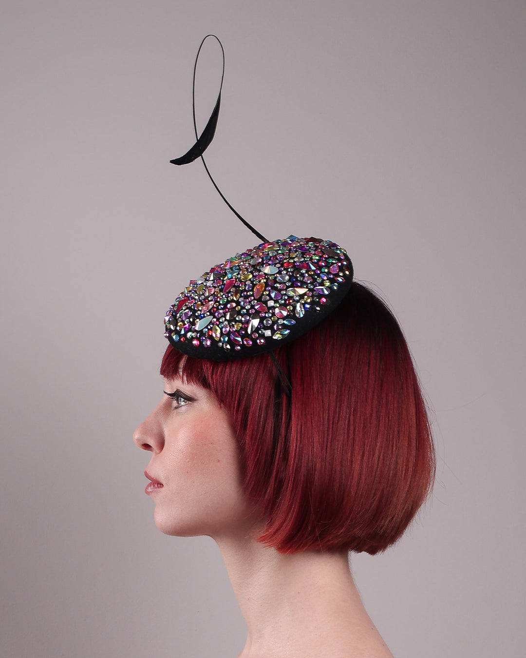 The model showcases the left side close up of the Celestine pillbox fascinator. Its structured, button-shaped black base is drenched in a kaleidoscope of iridescent crystals—pink, aqua, violet, and gold, among others—each catching the light in motion. Crowned with a sweeping black quill that curves like a clef in mid-air. Designed by Merve Bayindir.