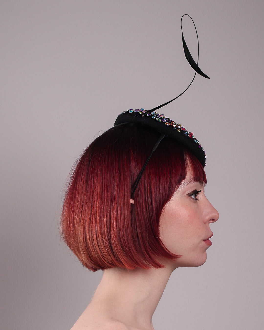 The model showcases the right side close up of the Celestine pillbox fascinator. Its structured, button-shaped black base is drenched in a kaleidoscope of iridescent crystals—pink, aqua, violet, and gold, among others—each catching the light in motion. Crowned with a sweeping black quill that curves like a clef in mid-air. Designed by Merve Bayindir.