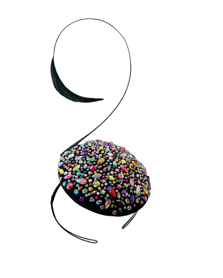 Still life picture of the Celestine pillbox fascinator. Its structured, button-shaped black base is drenched in a kaleidoscope of iridescent crystals—pink, aqua, violet, and gold, among others—each catching the light in motion. Crowned with a sweeping black quill that curves like a clef in mid-air. Designed by Merve Bayindir.