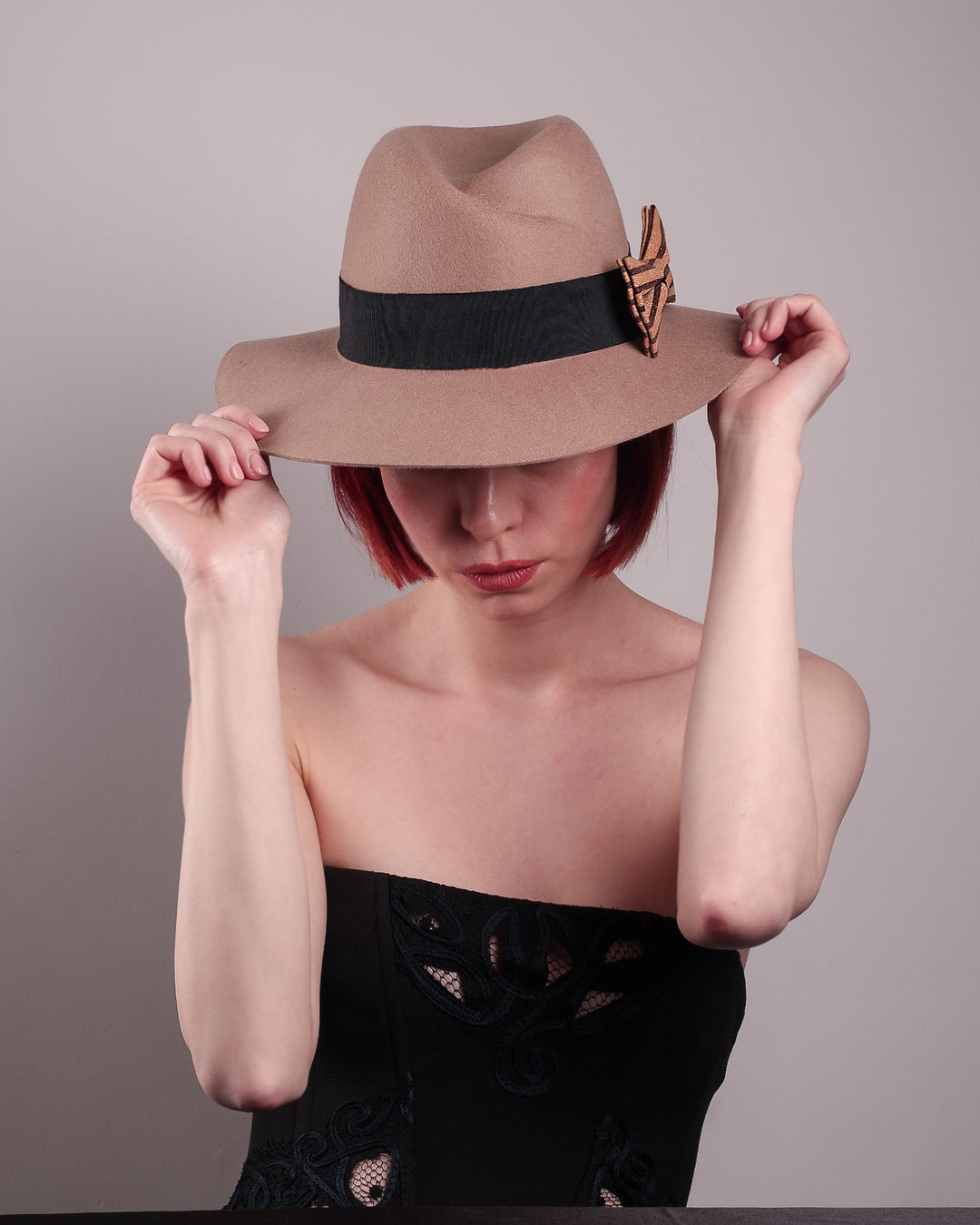 The model is showcasing the front from the top angle of the Celine, a classic light camel coloured fedora hat featuring a black stripe bow detail. Crafted from smooth, light camel felt with a gently pinched crown, this fedora is decorated with a sculptural bow, set off against a deep black grosgrain band. Designed by Merve Bayindir.