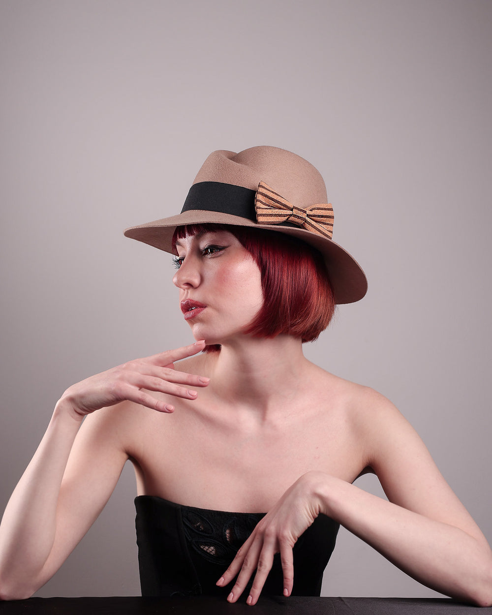 The model is showcasing the left low angle of the Celine, a classic light camel coloured fedora hat featuring a black stripe bow detail. Crafted from smooth, light camel felt with a gently pinched crown, this fedora is decorated with a sculptural bow, set off against a deep black grosgrain band. Designed by Merve Bayindir.