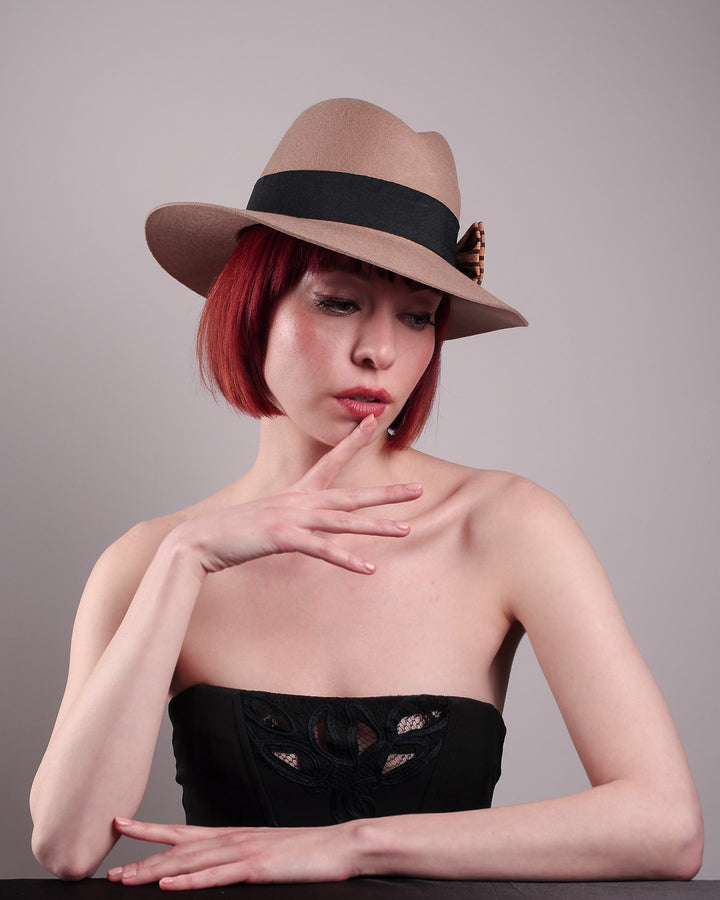 The model is showcasing the front right side of the Celine, a classic light camel coloured fedora hat featuring a black stripe bow detail. Crafted from smooth, light camel felt with a gently pinched crown, this fedora is decorated with a sculptural bow, set off against a deep black grosgrain band. Designed by Merve Bayindir.