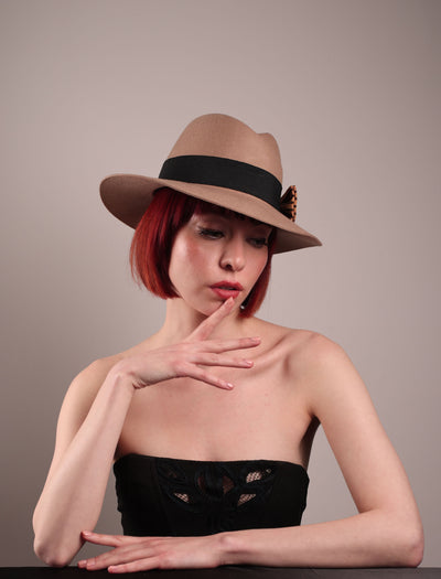 The model is showcasing the front right side of the Celine, a classic light camel coloured fedora hat featuring a black stripe bow detail. Crafted from smooth, light camel felt with a gently pinched crown, this fedora is decorated with a sculptural bow, set off against a deep black grosgrain band. Designed by Merve Bayindir.