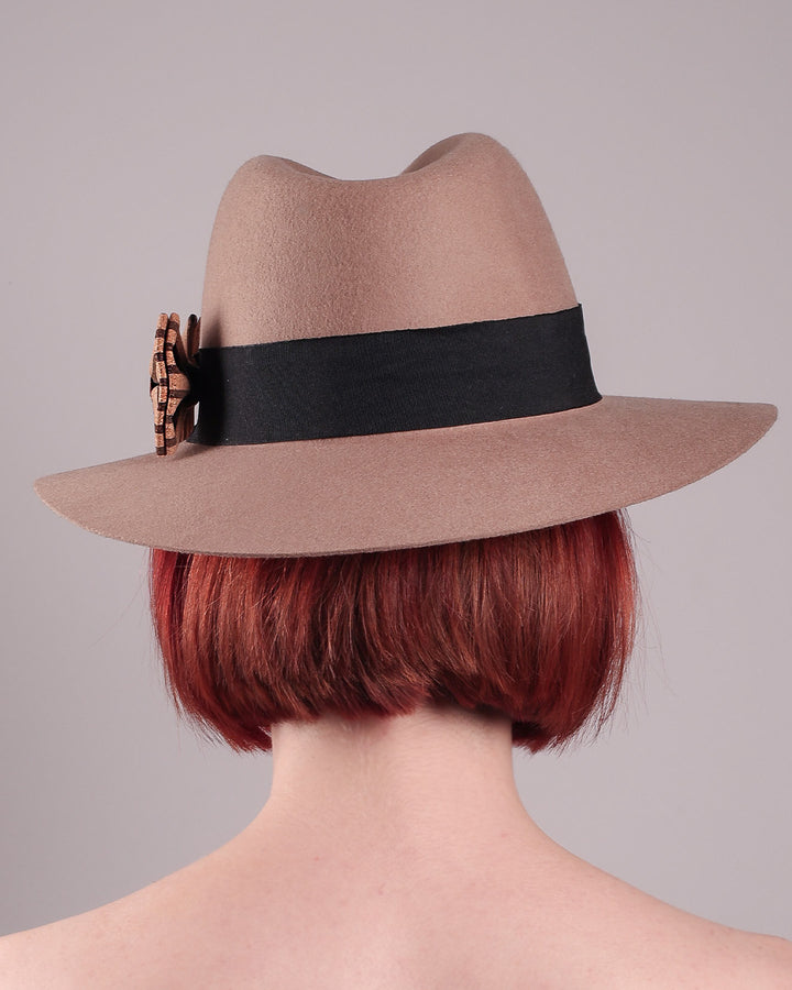 The model is showcasing the back of the Celine, a classic light camel coloured fedora hat featuring a black stripe bow detail. Crafted from smooth, light camel felt with a gently pinched crown, this fedora is decorated with a sculptural bow, set off against a deep black grosgrain band. Designed by Merve Bayindir.
