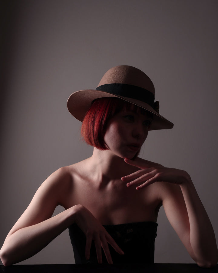 In the editorial photo with dark lighting the model is showcasing the front right side of the Celine, a classic light camel coloured fedora hat featuring a black stripe bow detail. Crafted from smooth, light camel felt with a gently pinched crown, this fedora is decorated with a sculptural bow, set off against a deep black grosgrain band. Designed by Merve Bayindir.