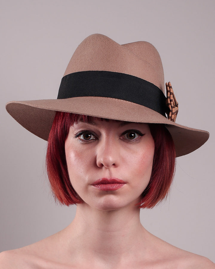 The model is showcasing the front close up of the Celine, a classic light camel coloured fedora hat featuring a black stripe bow detail. Crafted from smooth, light camel felt with a gently pinched crown, this fedora is decorated with a sculptural bow, set off against a deep black grosgrain band. Designed by Merve Bayindir.