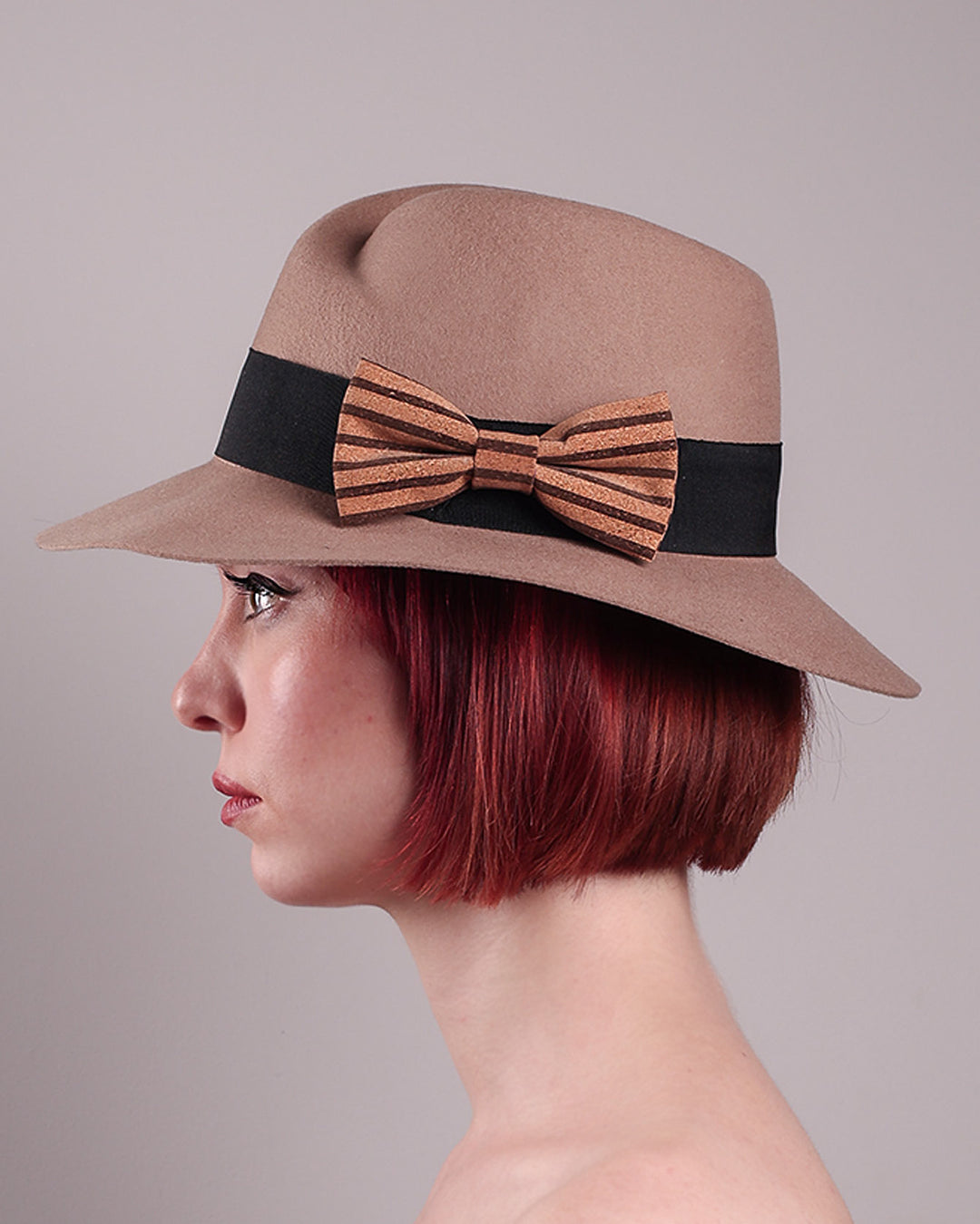 The model is showcasing the left side close up of the Celine, a classic light camel coloured fedora hat featuring a black stripe bow detail. Crafted from smooth, light camel felt with a gently pinched crown, this fedora is decorated with a sculptural bow, set off against a deep black grosgrain band. Designed by Merve Bayindir.