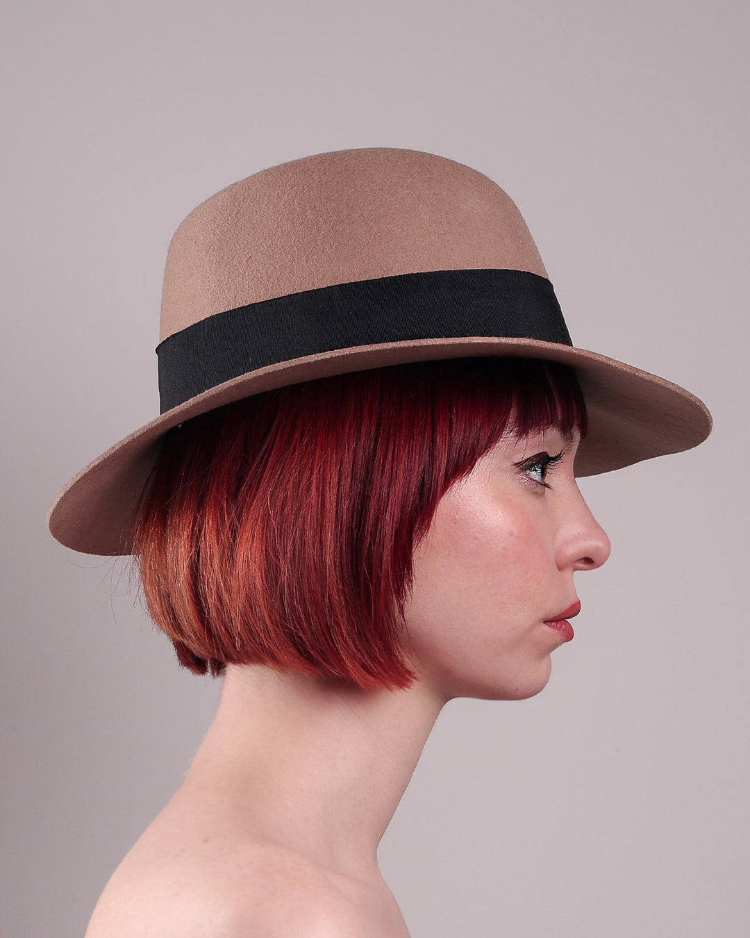The model is showcasing the right side close up of the Celine, a classic light camel coloured fedora hat featuring a black stripe bow detail. Crafted from smooth, light camel felt with a gently pinched crown, this fedora is decorated with a sculptural bow, set off against a deep black grosgrain band. Designed by Merve Bayindir.