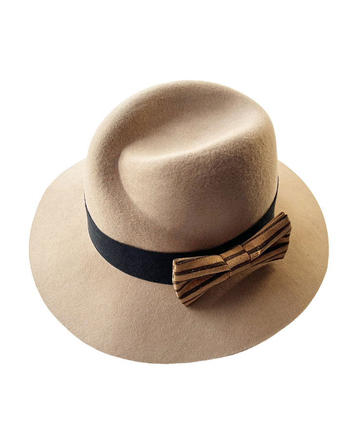 Still life picture of the Celine, a classic light camel coloured fedora hat featuring a black stripe bow detail. Crafted from smooth, light camel felt with a gently pinched crown, this fedora is decorated with a sculptural bow, set off against a deep black grosgrain band. Designed by Merve Bayindir.