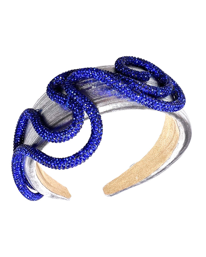 Light silver headband with a faux leather base, wrapped in bold looping cobalt blue crystal-embellished cords, creating a sculptural, high-shine statement design. Designed and made by Merve Bayindir Millinery.
