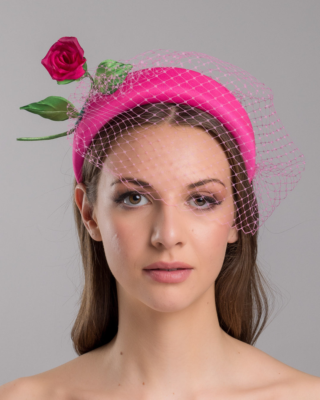 Model is show casing front close up of pink headband features soft, vibrant pink fabric that wraps comfortably around a sturdy band. Adorning one side of the headband is a beautifully crafted pink rose with lush green leaves. The rose is complemented by a delicate pink mesh veil that drapes gracefully over the headband. Made by Merve Bayindir Millinery.