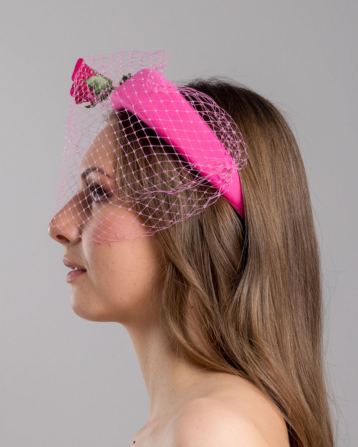 Model is show casing left side close up of pink headband features soft, vibrant pink fabric that wraps comfortably around a sturdy band. Adorning one side of the headband is a beautifully crafted pink rose with lush green leaves. The rose is complemented by a delicate pink mesh veil that drapes gracefully over the headband. Made by Merve Bayindir Millinery.