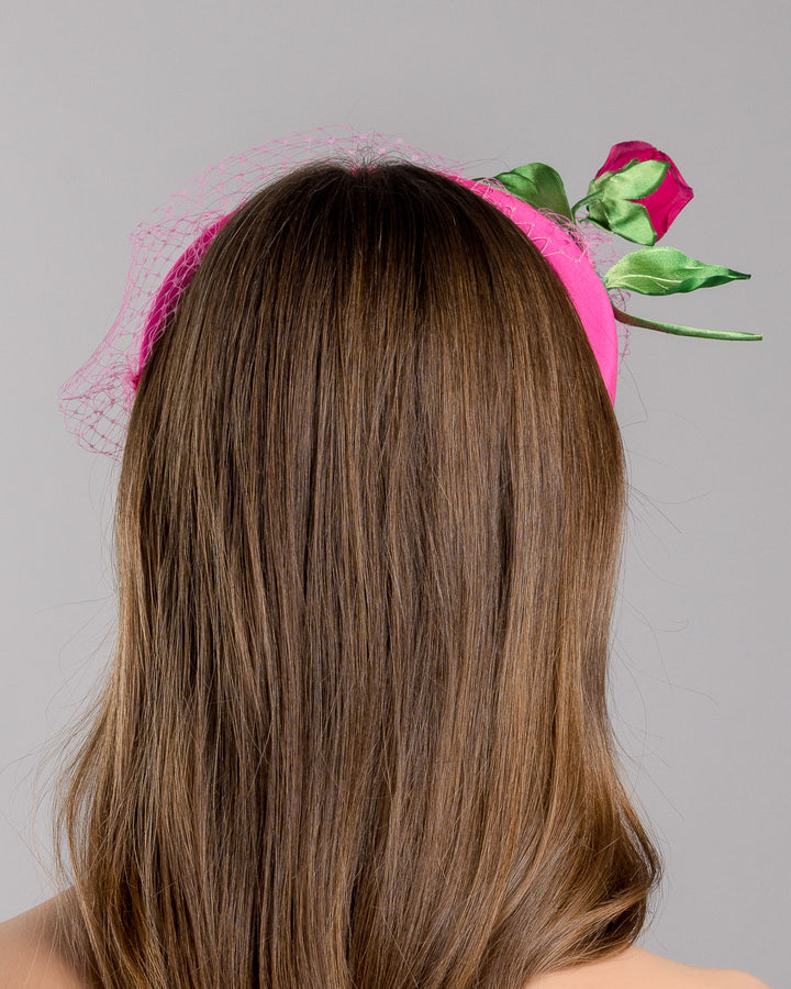 Model is show casing back side close up of pink headband features soft, vibrant pink fabric that wraps comfortably around a sturdy band. Adorning one side of the headband is a beautifully crafted pink rose with lush green leaves. The rose is complemented by a delicate pink mesh veil that drapes gracefully over the headband. Made by Merve Bayindir Millinery.