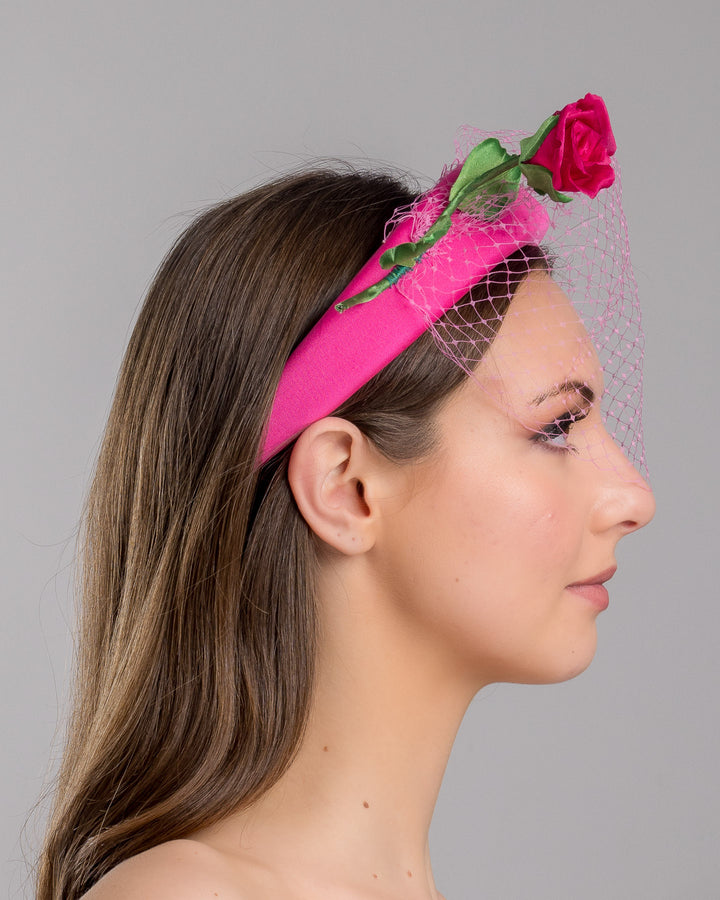 Model is show casing right side close up of pink headband features soft, vibrant pink fabric that wraps comfortably around a sturdy band. Adorning one side of the headband is a beautifully crafted pink rose with lush green leaves. The rose is complemented by a delicate pink mesh veil that drapes gracefully over the headband. Made by Merve Bayindir Millinery.