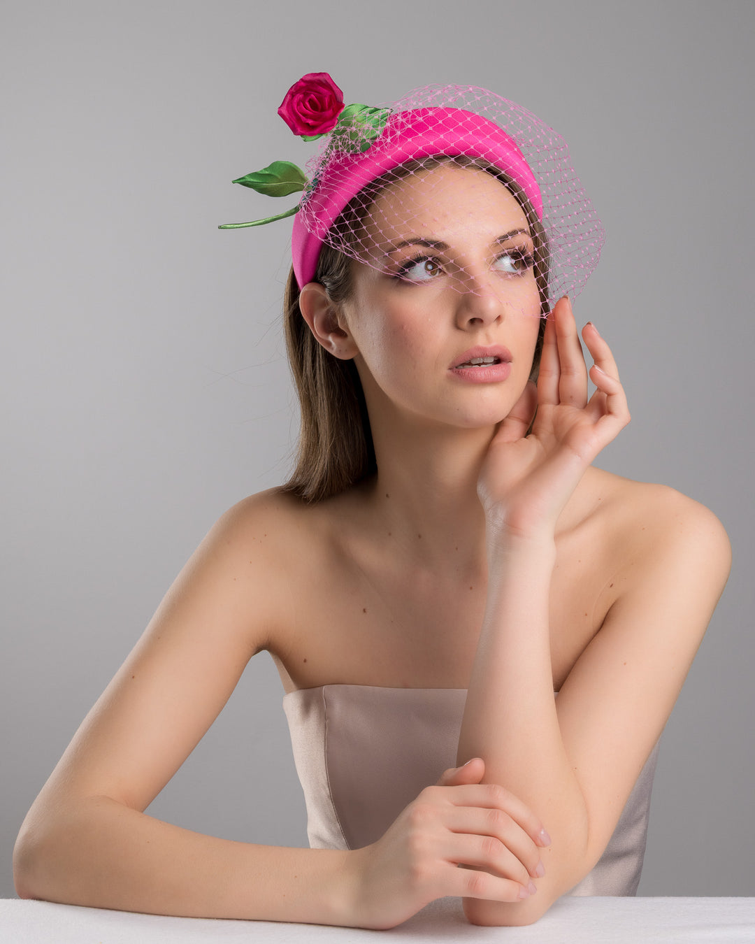 Female model is posing wearing Charleston pink headband features soft, vibrant pink fabric that wraps comfortably around a sturdy band. Adorning one side of the headband is a beautifully crafted pink rose with lush green leaves. The rose is complemented by a delicate pink mesh veil that drapes gracefully over the headband. Made by Merve Bayindir Millinery.