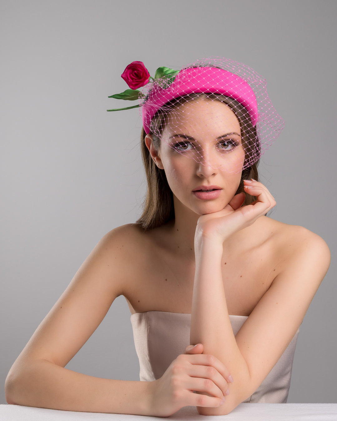 Female model is posing wearing Charleston pink headband features soft, vibrant pink fabric that wraps comfortably around a sturdy band. Adorning one side of the headband is a beautifully crafted pink rose with lush green leaves. The rose is complemented by a delicate pink mesh veil that drapes gracefully over the headband. Made by Merve Bayindir Millinery.