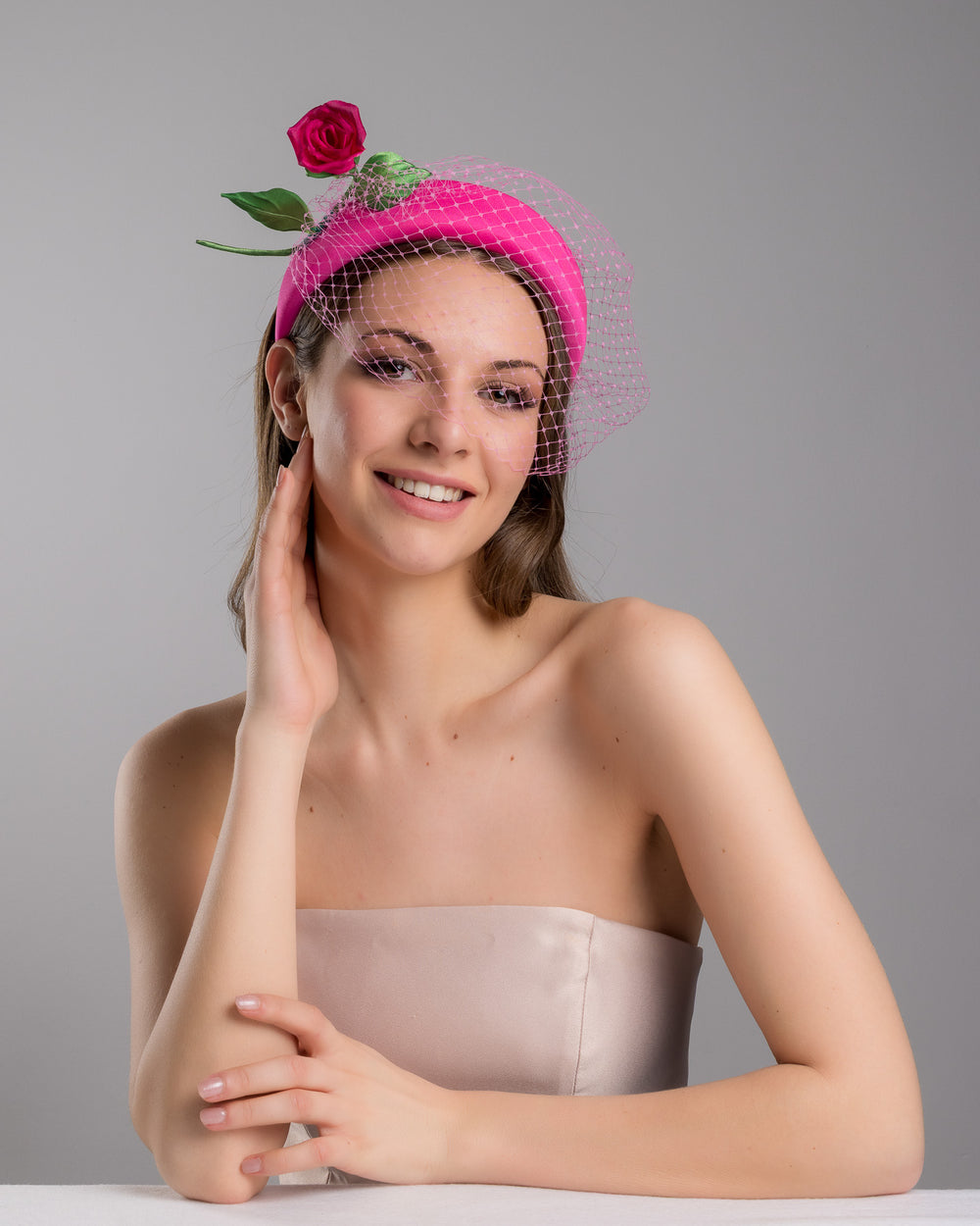 Female model is posing wearing Charleston pink headband features soft, vibrant pink fabric that wraps comfortably around a sturdy band. Adorning one side of the headband is a beautifully crafted pink rose with lush green leaves. The rose is complemented by a delicate pink mesh veil that drapes gracefully over the headband. Made by Merve Bayindir Millinery.