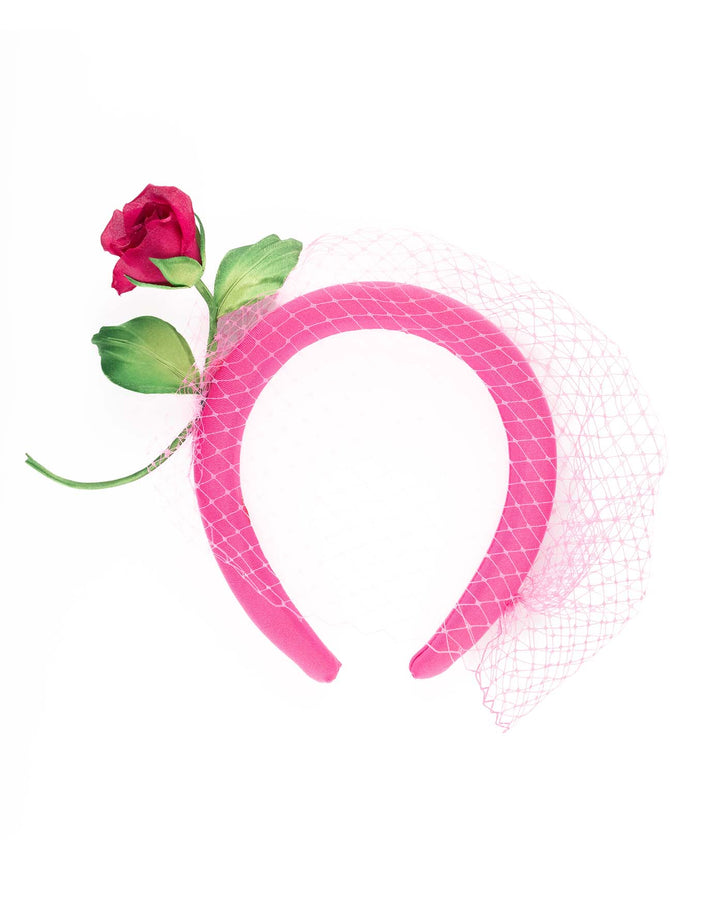 The pink headband features soft, vibrant pink fabric that wraps comfortably around a sturdy band. Adorning one side of the headband is a beautifully crafted pink rose with lush green leaves. The rose is complemented by a delicate pink mesh veil that drapes gracefully over the headband. Made by Merve Bayindir Millinery.