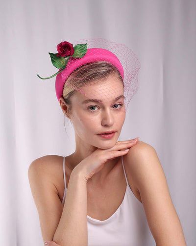 Model is wearing a pink headband features soft, vibrant pink fabric that wraps comfortably around a sturdy band. Adorning one side of the headband is a beautifully crafted pink rose with lush green leaves. The rose is complemented by a delicate pink mesh veil that drapes gracefully over the headband