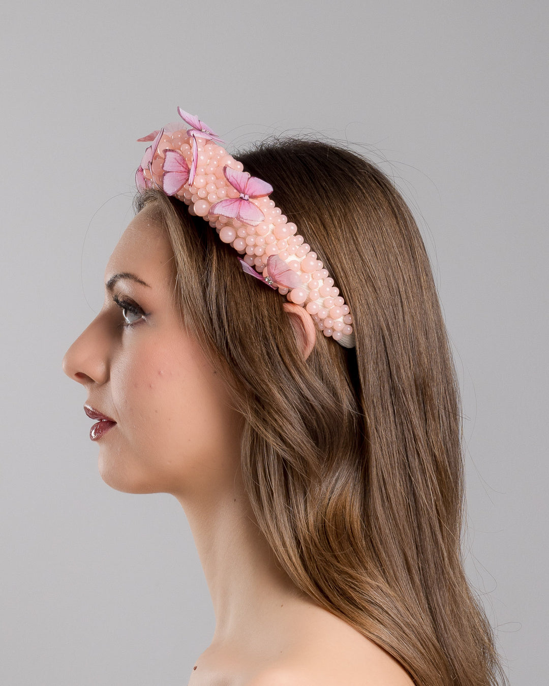 Model is showcasing the left side close up of high headband decorated with different sized mat peach beads and scattered pink organza butterflies with crystal centres. Made by Merve Bayindir millinery.