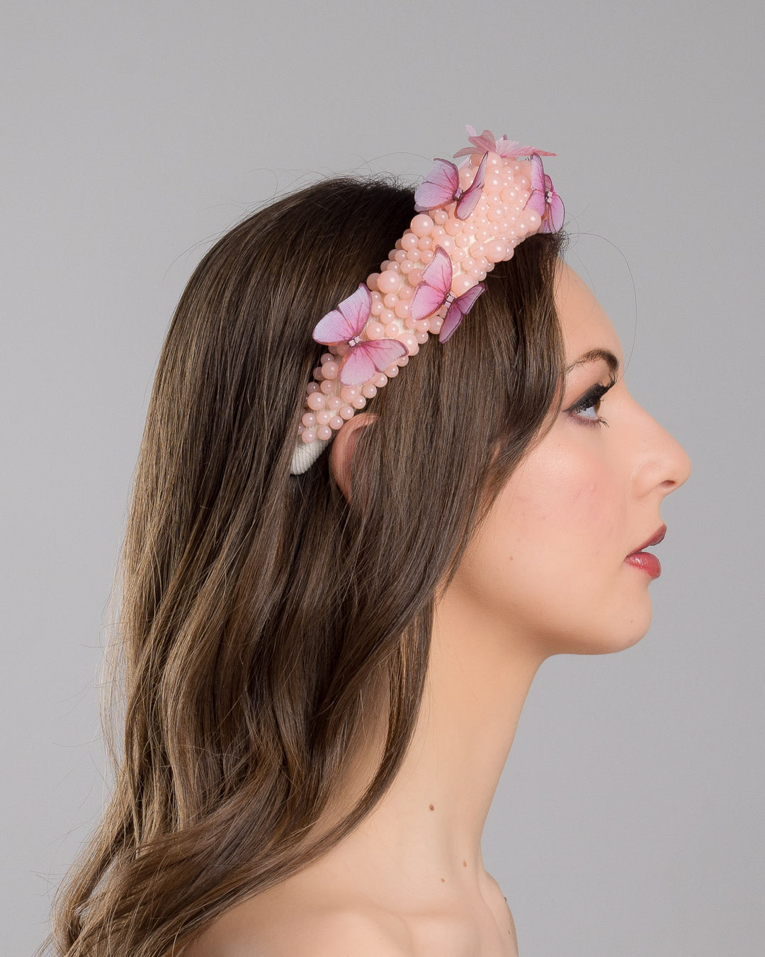 Model is showcasing the right side close up of high headband decorated with different sized mat peach beads and scattered pink organza butterflies with crystal centres. Made by Merve Bayindir millinery.
