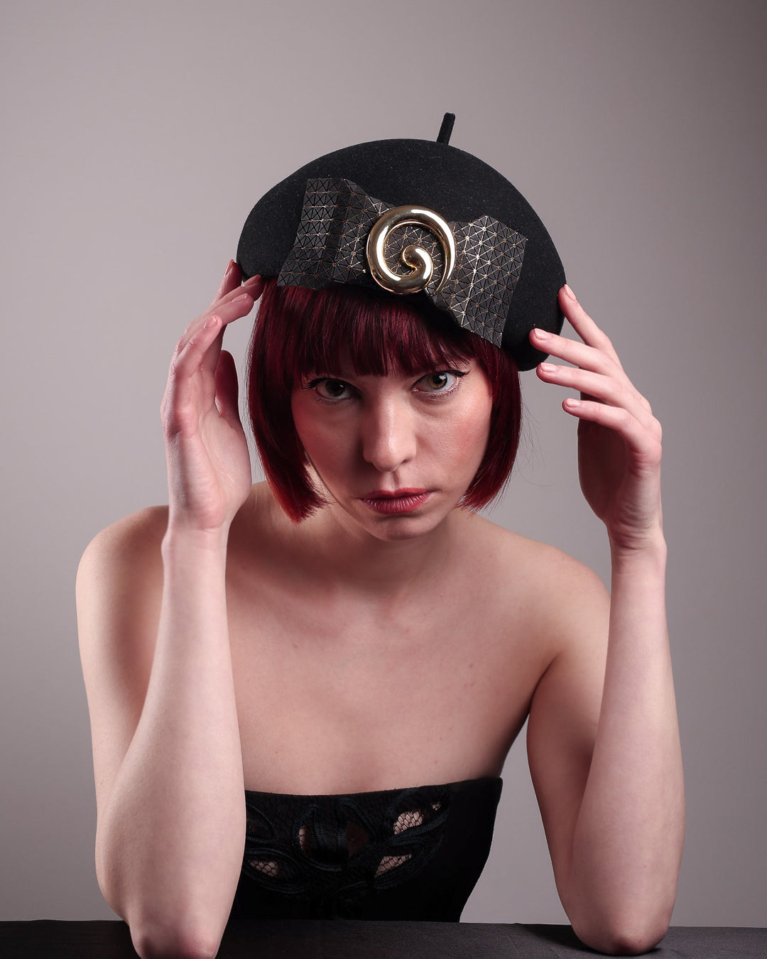 The model showcases the front view of the black dome-shaped, round-structured Clarisse wool felt pillbox beret. This is an embellished, textured piece with a gold metallic-based black faux leather bow tied in the centre, featuring a sculpted gold spiral. Designed by Merve Bayindir.