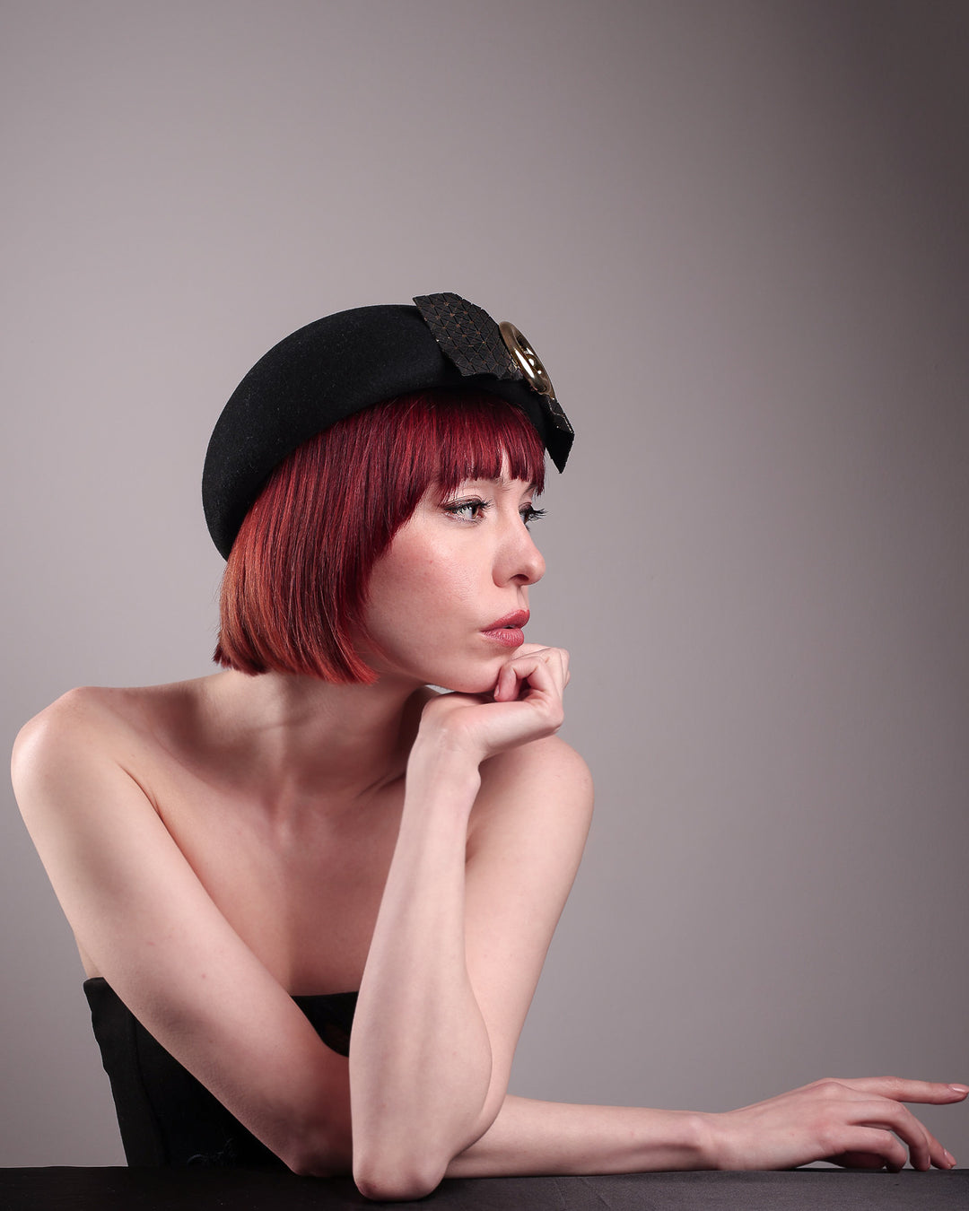 The model showcases the right side view of the black dome-shaped, round-structured Clarisse wool felt pillbox beret. This is an embellished, textured piece with a gold metallic-based black faux leather bow tied in the centre, featuring a sculpted gold spiral. Designed by Merve Bayindir.