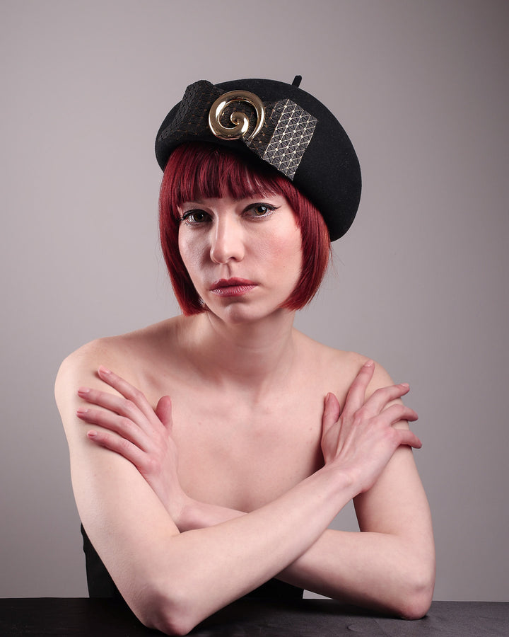 The model showcases the left angle of the black dome-shaped, round-structured Clarisse wool felt pillbox beret. This is an embellished, textured piece with a gold metallic-based black faux leather bow tied in the centre, featuring a sculpted gold spiral. Designed by Merve Bayindir.