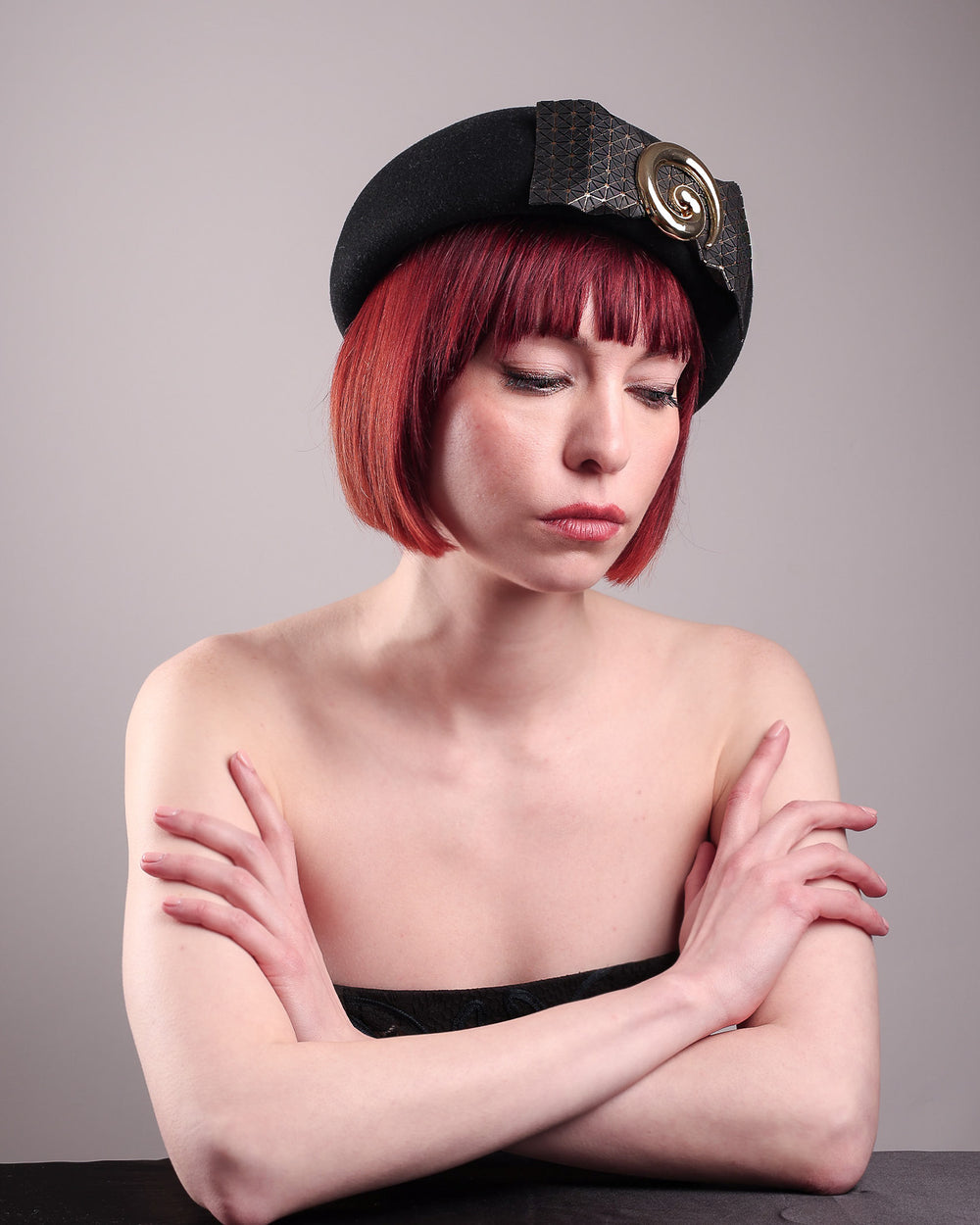 The model showcases the right bottom angle view of the black dome-shaped, round-structured Clarisse wool felt pillbox beret. This is an embellished, textured piece with a gold metallic-based black faux leather bow tied in the centre, featuring a sculpted gold spiral. Designed by Merve Bayindir.