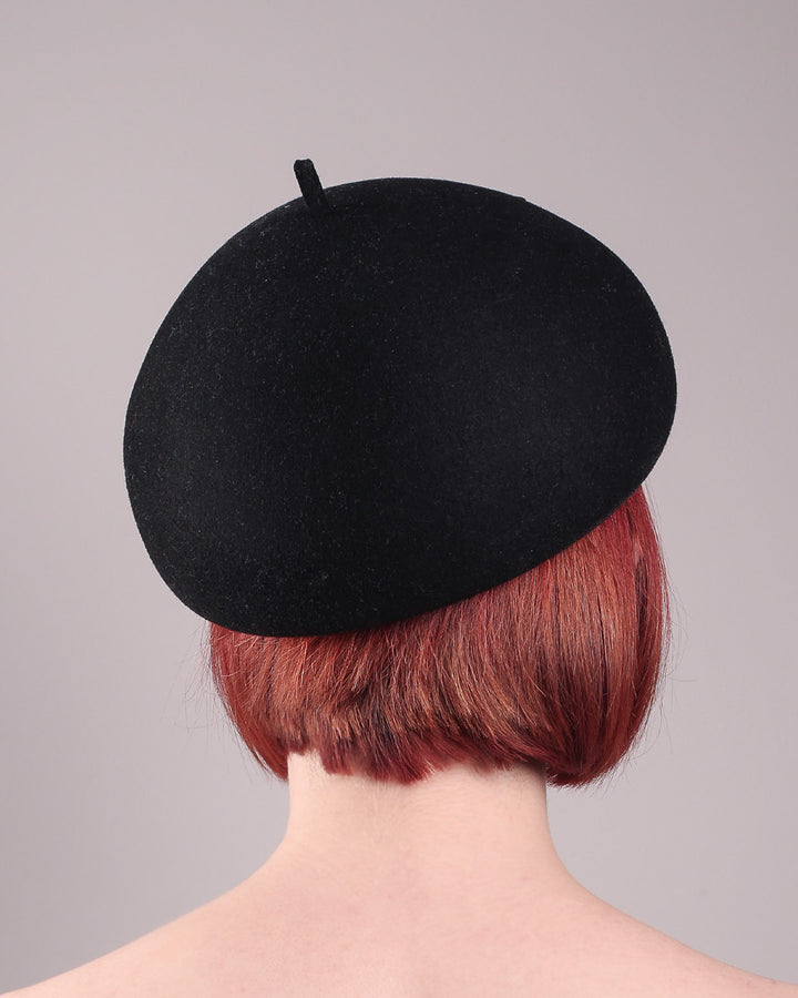 The model showcases the back of the black dome-shaped, round-structured Clarisse wool felt pillbox beret. This is an embellished, textured piece with a gold metallic-based black faux leather bow tied in the centre, featuring a sculpted gold spiral. Designed by Merve Bayindir.
