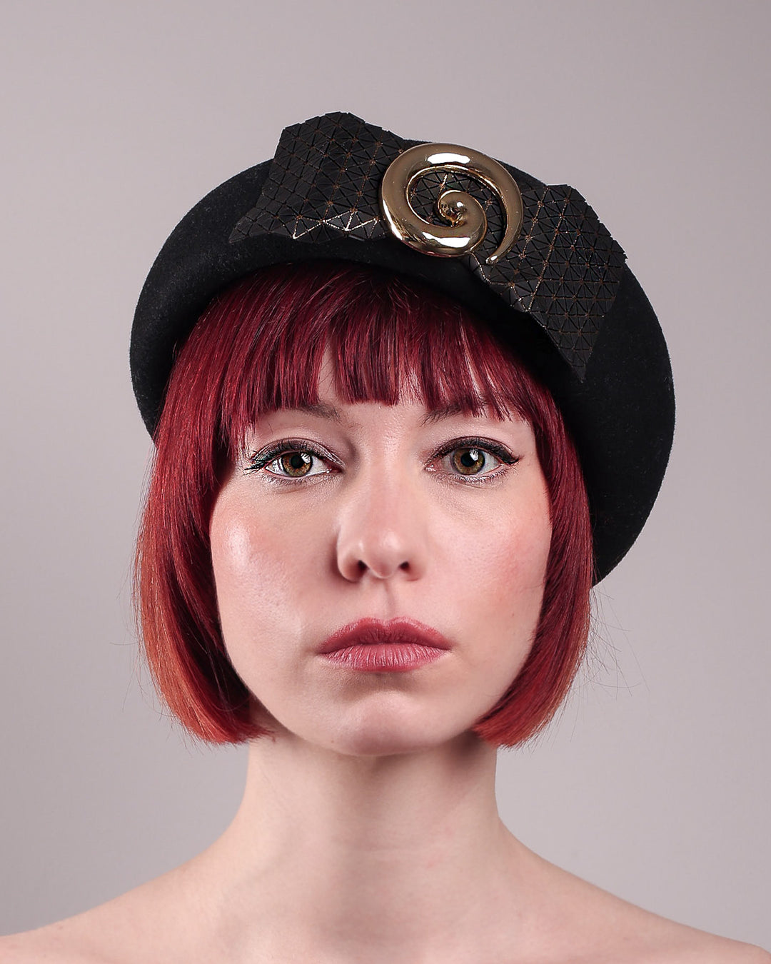 The model showcases the front close up of the black dome-shaped, round-structured Clarisse wool felt pillbox beret. This is an embellished, textured piece with a gold metallic-based black faux leather bow tied in the centre, featuring a sculpted gold spiral. Designed by Merve Bayindir.
