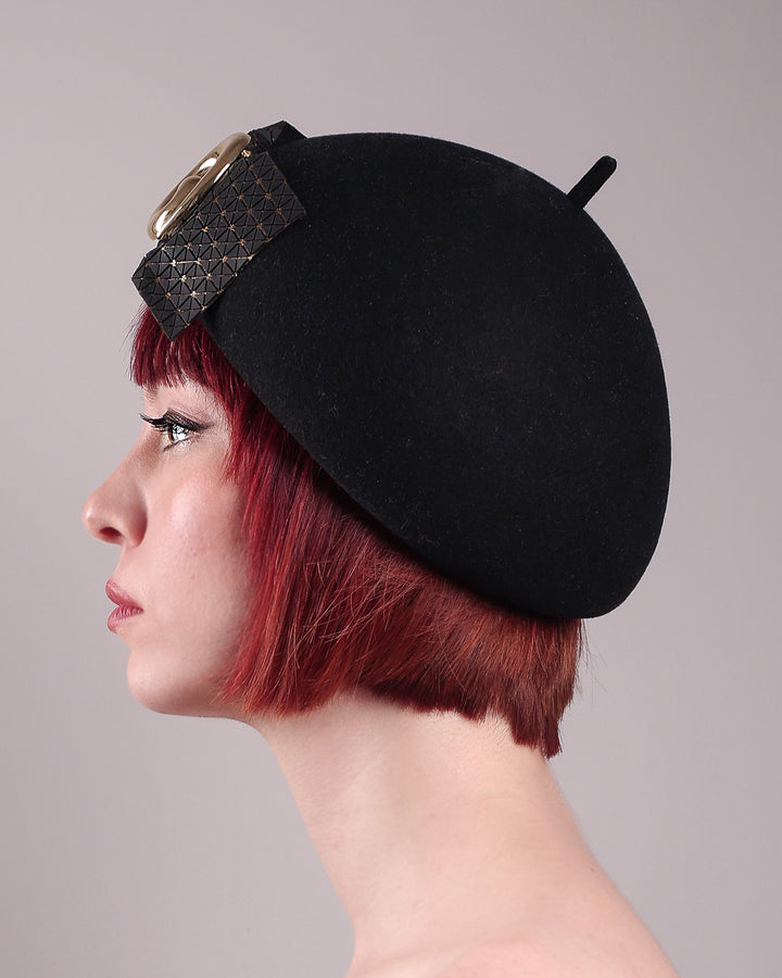 The model showcases the left side close up of the black dome-shaped, round-structured Clarisse wool felt pillbox beret. This is an embellished, textured piece with a gold metallic-based black faux leather bow tied in the centre, featuring a sculpted gold spiral. Designed by Merve Bayindir.
