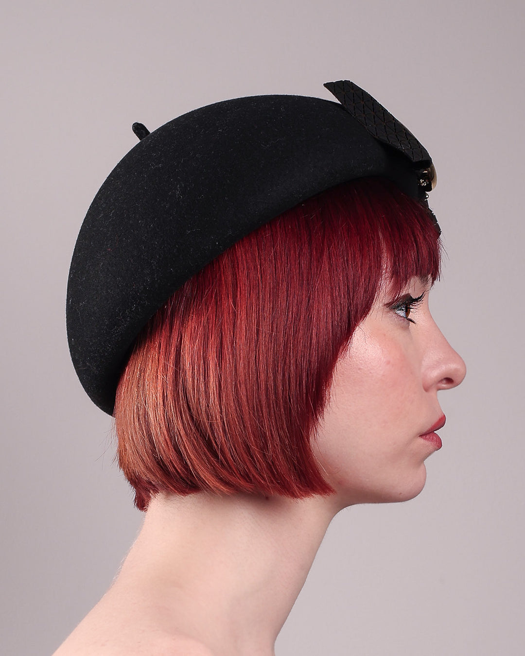 The model showcases the right side close up of the black dome-shaped, round-structured Clarisse wool felt pillbox beret. This is an embellished, textured piece with a gold metallic-based black faux leather bow tied in the centre, featuring a sculpted gold spiral. Designed by Merve Bayindir.