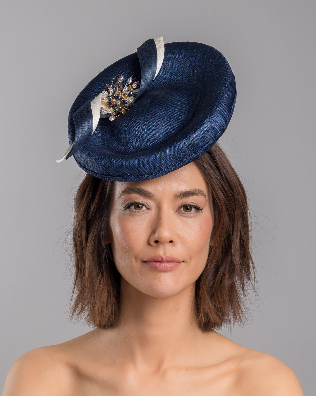 Model is showcasing the front side close up of oval navy fascinator with a structured oval shape sits high on the head. The design is accentuated by graceful navy and white swirls that loop stylishly across the brim, creating a dynamic and eye-catching detail. At the centre of the ribbon, a stunning cluster of crystal beads in various sizes and shades forms a beautiful motif. Designed and made by Merve Bayindir millinery.