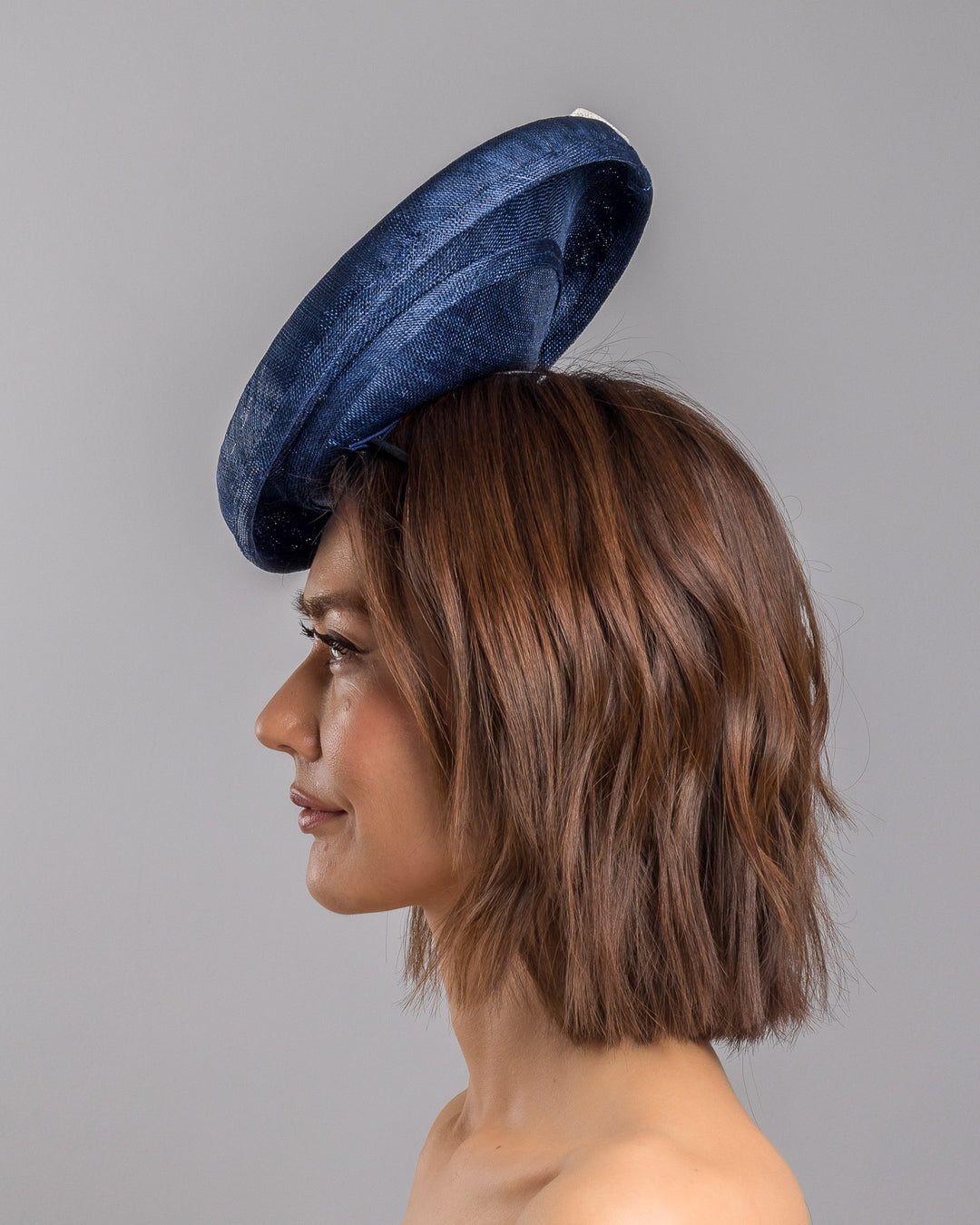 Model is showcasing the left side close up of oval navy fascinator with a structured oval shape sits high on the head. The design is accentuated by graceful navy and white swirls that loop stylishly across the brim, creating a dynamic and eye-catching detail. At the centre of the ribbon, a stunning cluster of crystal beads in various sizes and shades forms a beautiful motif. Designed and made by Merve Bayindir millinery.