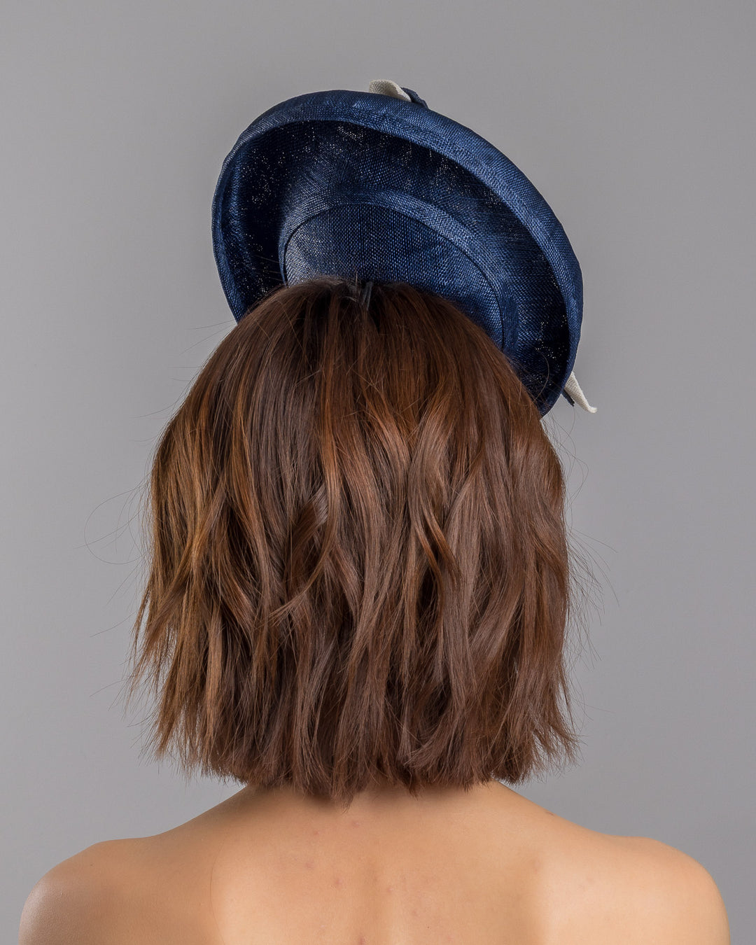 Model is showcasing the back side close up of oval navy fascinator with a structured oval shape sits high on the head. The design is accentuated by graceful navy and white swirls that loop stylishly across the brim, creating a dynamic and eye-catching detail. At the centre of the ribbon, a stunning cluster of crystal beads in various sizes and shades forms a beautiful motif. Designed and made by Merve Bayindir millinery.