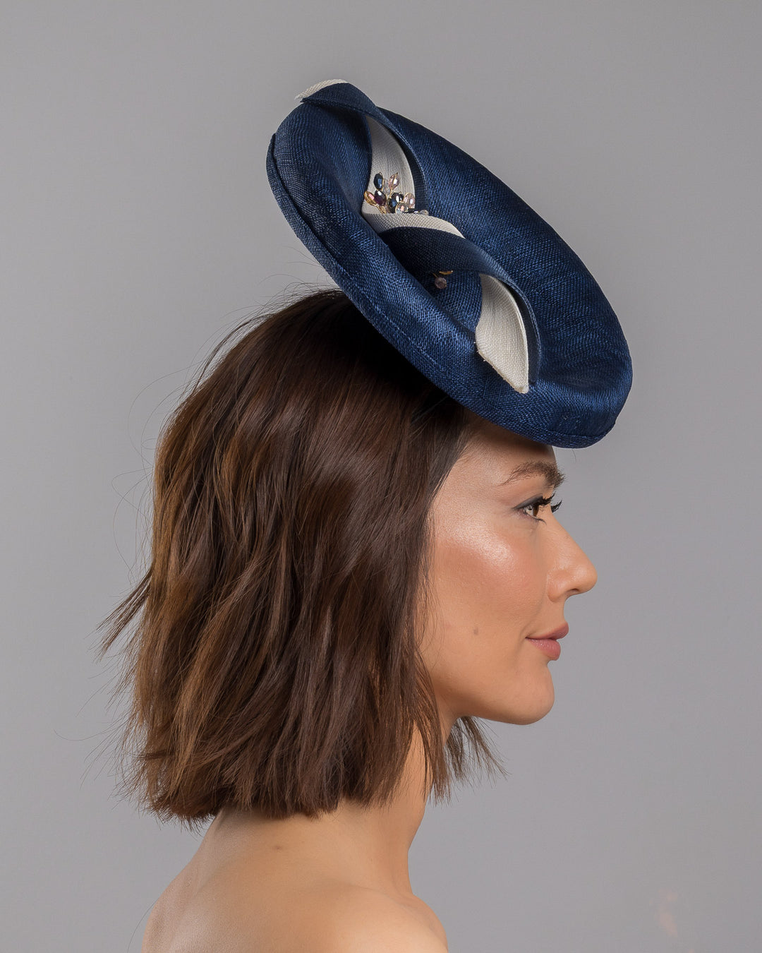 Model is showcasing the right side close up of oval navy fascinator with a structured oval shape sits high on the head. The design is accentuated by graceful navy and white swirls that loop stylishly across the brim, creating a dynamic and eye-catching detail. At the centre of the ribbon, a stunning cluster of crystal beads in various sizes and shades forms a beautiful motif. Designed and made by Merve Bayindir millinery.