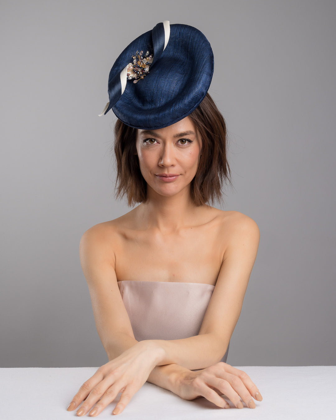 Model is posing wearing Clay an oval navy fascinator with a structured oval shape sits high on the head. The design is accentuated by graceful navy and white swirls that loop stylishly across the brim, creating a dynamic and eye-catching detail. At the centre of the ribbon, a stunning cluster of crystal beads in various sizes and shades forms a beautiful motif. Designed and made by Merve Bayindir millinery.