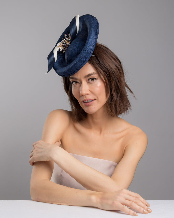 Model is posing wearing Clay an oval navy fascinator with a structured oval shape sits high on the head. The design is accentuated by graceful navy and white swirls that loop stylishly across the brim, creating a dynamic and eye-catching detail. At the centre of the ribbon, a stunning cluster of crystal beads in various sizes and shades forms a beautiful motif. Designed and made by Merve Bayindir millinery.