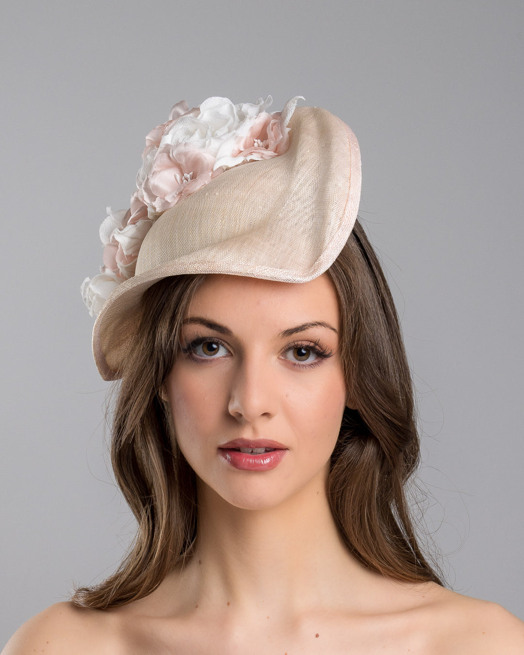 The model is showcasing the front side close up of the delicate and elegant pink fascinator hat featuring a soft blush pink fabric with a subtle shimmer. The hat has a structured, crescent shape that arches, providing a flattering and sophisticated silhouette. Adorning the top of the hat is a beautiful arrangement of handmade flowers in soft pink and white hues. Designed and made by Merve Bayindir Millinery.
