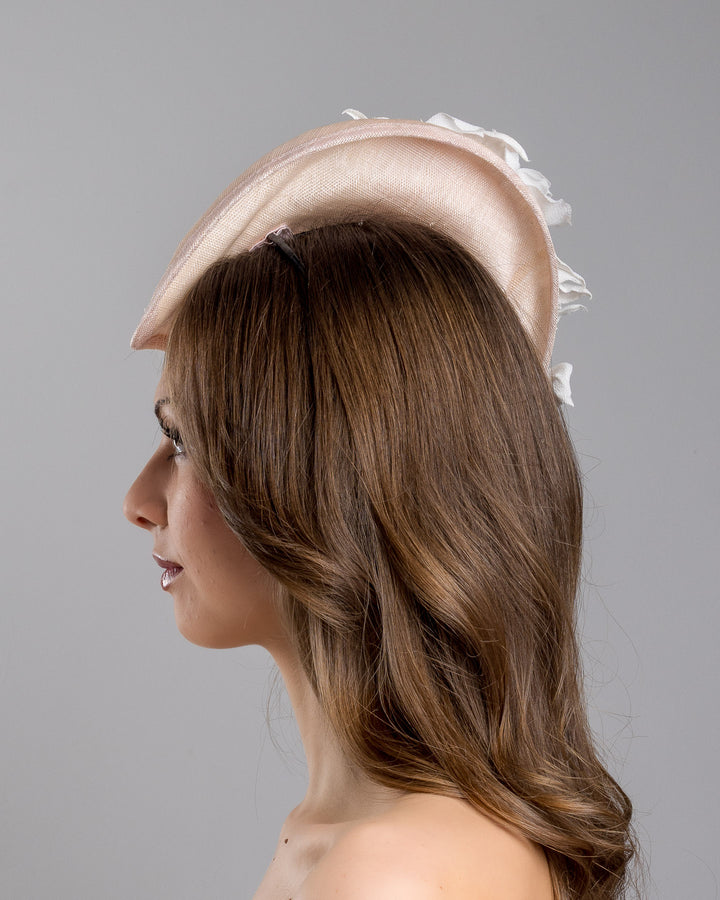 The model is showcasing the left side close up of the delicate and elegant pink fascinator hat featuring a soft blush pink fabric with a subtle shimmer. The hat has a structured, crescent shape that arches, providing a flattering and sophisticated silhouette. Adorning the top of the hat is a beautiful arrangement of handmade flowers in soft pink and white hues. Designed and made by Merve Bayindir Millinery.
