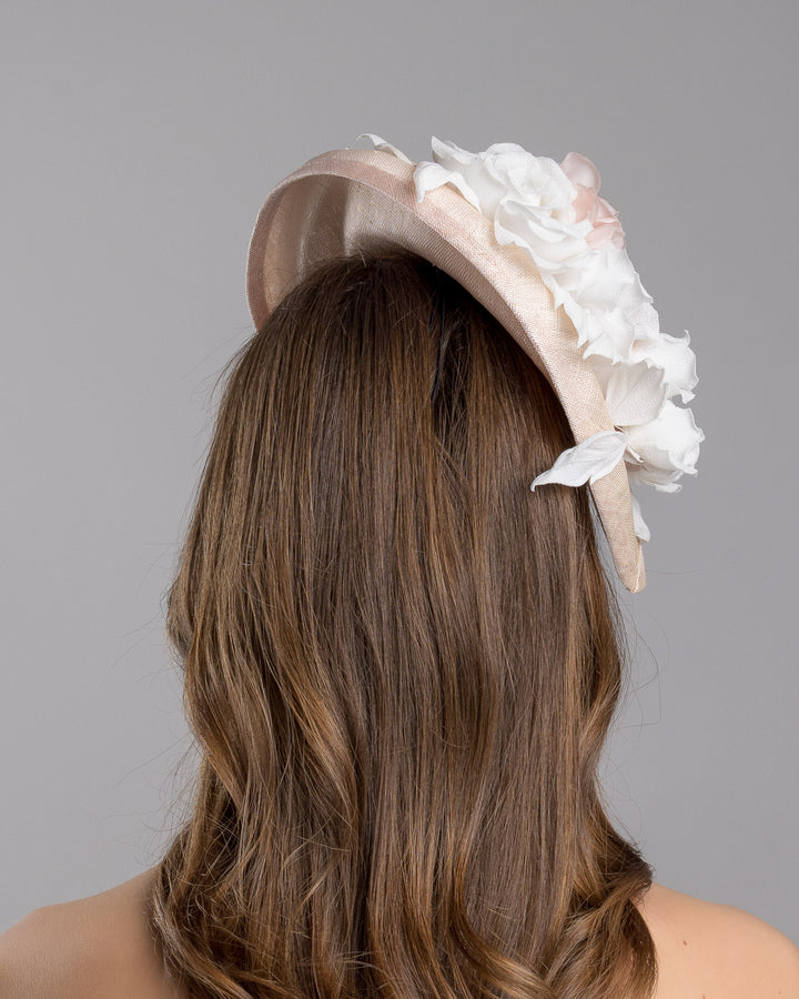The model is showcasing the back side close up of the delicate and elegant pink fascinator hat featuring a soft blush pink fabric with a subtle shimmer. The hat has a structured, crescent shape that arches, providing a flattering and sophisticated silhouette. Adorning the top of the hat is a beautiful arrangement of handmade flowers in soft pink and white hues. Designed and made by Merve Bayindir Millinery.
