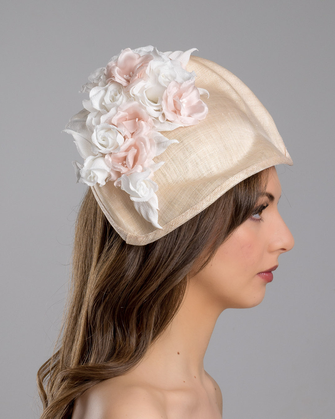 The model is showcasing the right side close up of the delicate and elegant pink fascinator hat featuring a soft blush pink fabric with a subtle shimmer. The hat has a structured, crescent shape that arches, providing a flattering and sophisticated silhouette. Adorning the top of the hat is a beautiful arrangement of handmade flowers in soft pink and white hues. Designed and made by Merve Bayindir Millinery.
