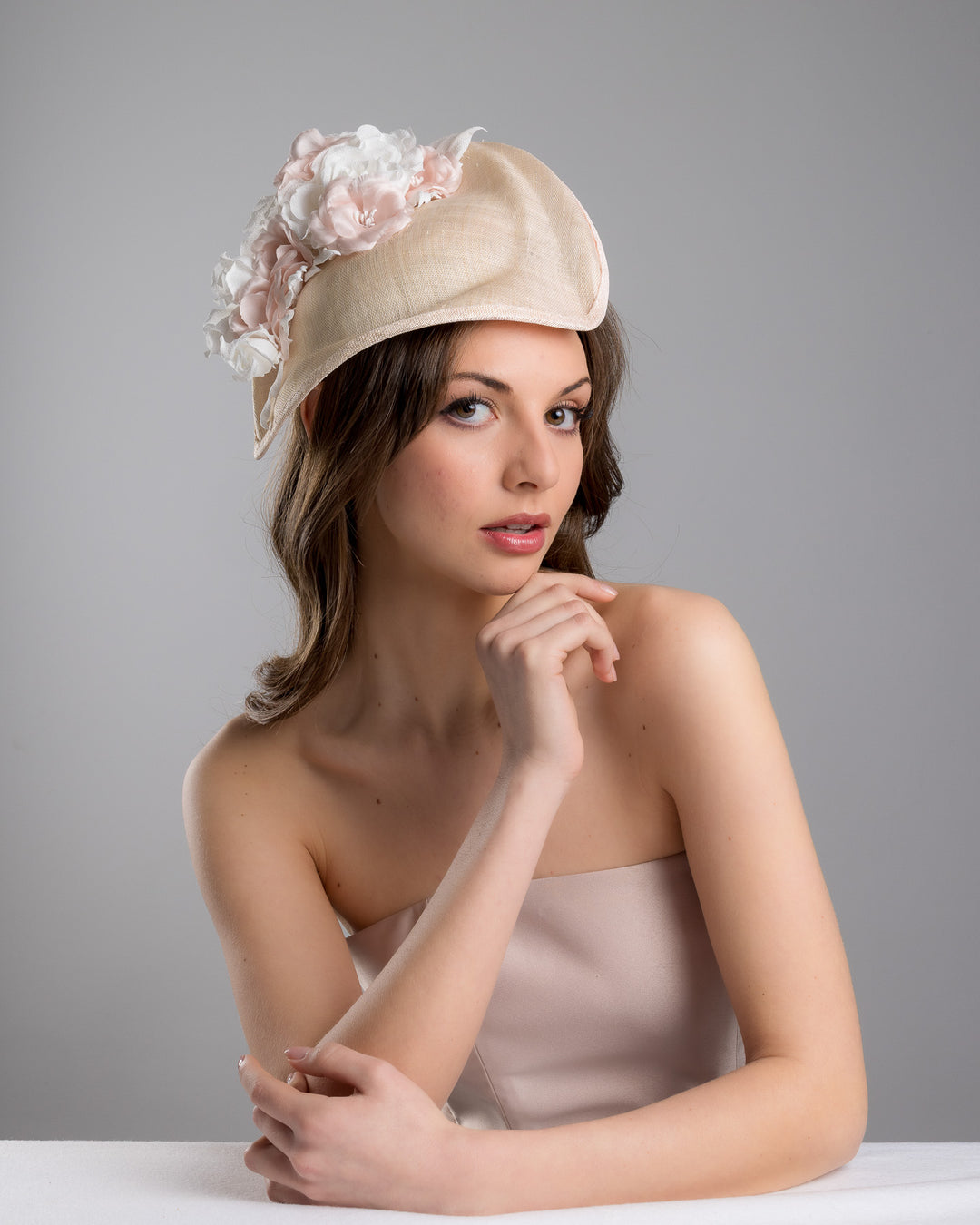The female model is posing, wearing Cleo, the delicate and elegant pink fascinator hat featuring a soft blush pink fabric with a subtle shimmer. The hat has a structured, crescent shape that arches, providing a flattering and sophisticated silhouette. Adorning the top of the hat is a beautiful arrangement of handmade flowers in soft pink and white hues. Designed and made by Merve Bayindir Millinery.
