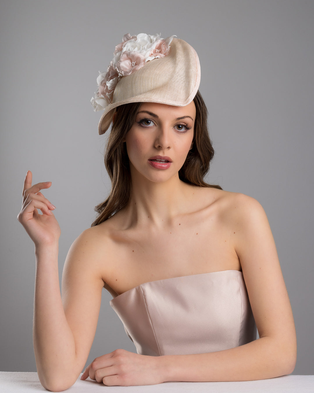 The female model is posing, wearing Cleo, the delicate and elegant pink fascinator hat featuring a soft blush pink fabric with a subtle shimmer. The hat has a structured, crescent shape that arches, providing a flattering and sophisticated silhouette. Adorning the top of the hat is a beautiful arrangement of handmade flowers in soft pink and white hues. Designed and made by Merve Bayindir Millinery.
