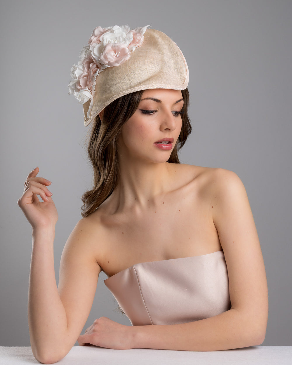 The female model is posing, wearing Cleo, the delicate and elegant pink fascinator hat featuring a soft blush pink fabric with a subtle shimmer. The hat has a structured, crescent shape that arches, providing a flattering and sophisticated silhouette. Adorning the top of the hat is a beautiful arrangement of handmade flowers in soft pink and white hues. Designed and made by Merve Bayindir Millinery.
