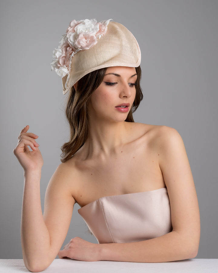 The female model is posing, wearing Cleo, the delicate and elegant pink fascinator hat featuring a soft blush pink fabric with a subtle shimmer. The hat has a structured, crescent shape that arches, providing a flattering and sophisticated silhouette. Adorning the top of the hat is a beautiful arrangement of handmade flowers in soft pink and white hues. Designed and made by Merve Bayindir Millinery.
